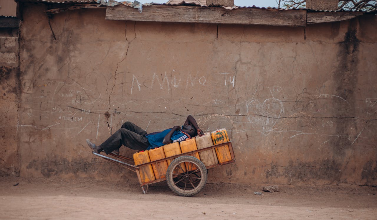 A man lies on a cart filled with containers beside a rustic wall in daylight.